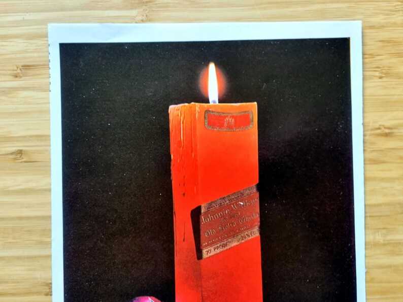 May include: A lit, rectangular red candle with a label that reads "Johnnie Walker Old Scotch Whisky". The candle is set against a black background. The flame is bright and yellow.