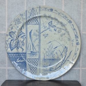 Peut inclure: Une assiette ronde de style ancien avec un motif nautique bleu et blanc. L'assiette présente un voilier, des motifs floraux et un paysage. Le bord est orné d'une délicate bordure florale. L'assiette est probablement en céramique.