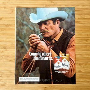 Könnte beinhalten: Vintage-Werbung mit einem Mann mit Cowboyhut, der eine Zigarette anzündet. Der Text lautet: „Komm dorthin, wo der Geschmack ist.“ Zwei Packungen Marlboro-Zigaretten sind ebenfalls zu sehen.