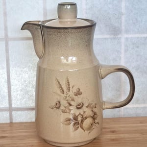 Könnte beinhalten: Eine beige Keramik-Kaffeekanne mit Deckel und Griff. Die Kanne hat ein Blumendesign in Braun- und Beigetönen. Der Rand und der Griff sind dunkler braun. Die Kanne steht auf einer Holzoberfläche.