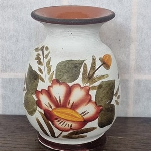 Puede incluir: Un jarrón de cerámica blanco con un borde marrón y un diseño floral. El jarrón presenta una gran flor roja con detalles amarillos y hojas verdes.