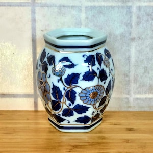 May include: A white and blue floral vase with a hexagonal base. The vase features a detailed design of blue flowers and leaves with gold accents. The rim is a light blue colour. The vase is on a wooden surface.