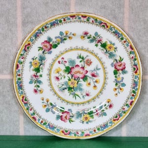 May include: A white porcelain plate with a floral design. The plate has a gold rim and a center design of pink, yellow, and green flowers. The border of the plate is decorated with a repeating pattern of blue, green, and yellow flowers.