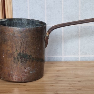 Puede incluir: Una cacerola de cobre vintage con un mango largo y delgado. La olla tiene forma cilíndrica con un borde ligeramente inclinado. El cobre tiene un aspecto envejecido y desgastado, con zonas de decoloración y pátina. El mango está unido al lateral de la olla.