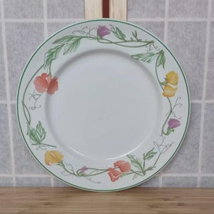 May include: White ceramic plate with a green border and floral design. The plate features a garland of colourful flowers in shades of orange, yellow, and purple, with green leaves. A decorative piece for the dining table.
