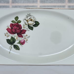 Puede incluir: Plato ovalado para servir de color crema con ribete dorado. El plato presenta un diseño floral de rosas rojas y blancas con hojas verdes. El plato tiene forma rectangular con bordes curvos.