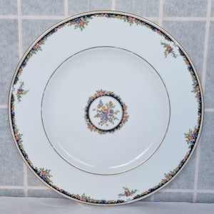 May include: A white porcelain plate with a gold rim and a black border decorated with floral patterns. The centre has a floral bouquet within a black circle. The plate is designed for serving food.