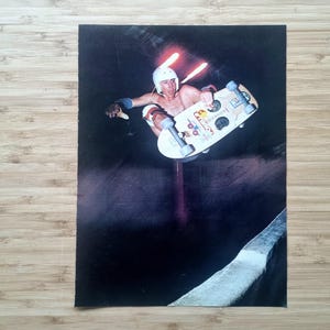 May include: Vintage photograph of a skateboarder mid-air, performing a trick. The skateboarder wears a white helmet, knee pads, and wrist guards. The skateboard has stickers. The background is dark with streaks of light.