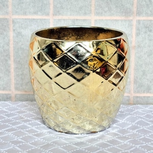以下が含まれることがあります： ダイヤモンド模様の金色の円筒形の花瓶。 花瓶は広い開口部とテクスチャ加工された表面を持っています。 花瓶は白い模様のテーブルクロスの上にあります。 インテリア。