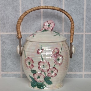Puede incluir: Jarra de cerámica color crema con tapa y asa trenzada. La jarra está decorada con diseños florales rosas y hojas verdes. El asa es de material marrón trenzado.