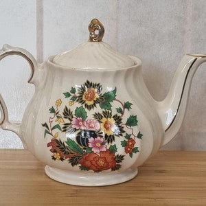 May include: A white teapot with a gold lid and floral design. The teapot has a round body and a curved spout. The floral design features pink, yellow, red, and blue flowers.