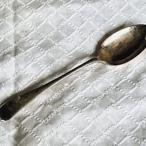 May include: A silver spoon with a long handle and an oval bowl. The spoon is resting on a white, textured surface with a diamond pattern. The spoon appears to be vintage or antique.