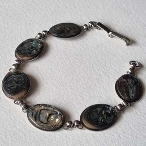 May include: Silver bracelet with oval abalone shell inlays. The iridescent shells are set in a silver metal frame and linked with small silver beads. The bracelet has a clasp closure. The jewellery is displayed on a white surface.