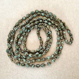 May include: A long necklace featuring oval teal beads with brown markings, linked with small gold-coloured beads. The necklace is arranged in a spiral, highlighting its length and design. A silver clasp secures the necklace. The necklace's length is visible.