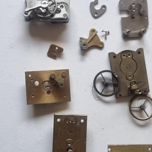 May include: An assortment of vintage watch parts, including gears, plates, and springs, in various shapes and sizes. The components are made of metal, with a mix of silver and brass tones. The arrangement suggests a disassembled timepiece.
