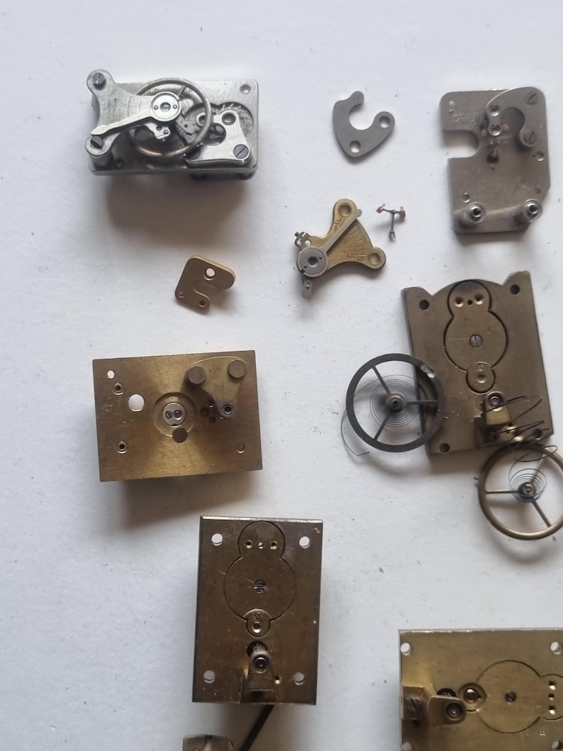 May include: An assortment of vintage watch parts, including gears, plates, and springs, in various shapes and sizes. The components are made of metal, with a mix of silver and brass tones. The arrangement suggests a disassembled timepiece.
