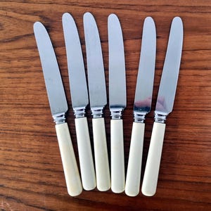 Può includere: Sei coltelli vintage con lame in acciaio inossidabile e manici color crema sono disposti su una superficie di legno. I coltelli sono distanziati in modo uniforme, mostrando il loro design classico. Le lame sono lucide e i manici lisci.