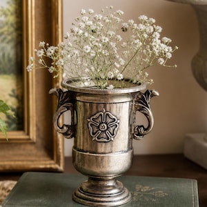 May include: A silver-coloured metal vase with a floral emblem and two handles, holding white baby's breath. The vase rests on a stack of books with gold lettering. A framed painting is in the background.