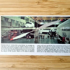 May include: Image of the Gossamer Albatross ultralight aircraft propeller. The propeller is white with red tips, and is in front of the aircraft. Text on the image reads "PROPELLER OF 'GOSSAMER ALBATROSS'". The aircraft is inside a hangar.