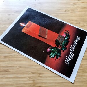 May include: A vintage Christmas card depicting a red candle with a lit flame, a sprig of holly, and the words "Happy Christmas" in white script. The background fades from black to red.