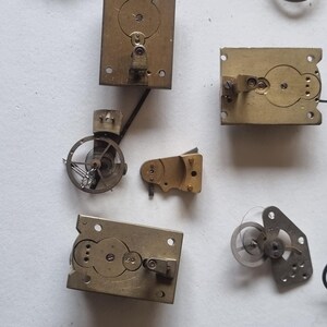 May include: A collection of vintage clock parts, including brass plates with circular cutouts, small gears, and springs. The components are arranged on a light-colored surface, showcasing the intricate details of the clock mechanisms.