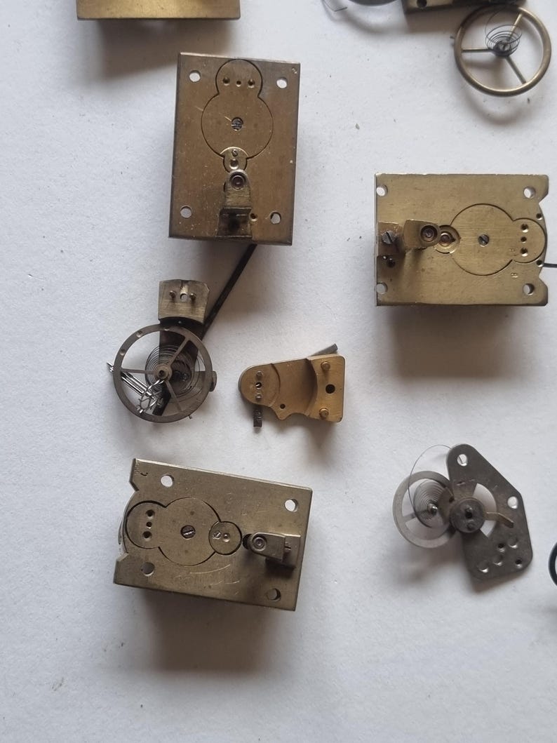 May include: A collection of vintage clock parts, including brass plates with circular cutouts, small gears, and springs. The components are arranged on a light-colored surface, showcasing the intricate details of the clock mechanisms.