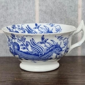 May include: A white teacup with a blue and white floral design featuring a winged dragon. The teacup has a handle and a rounded base.
