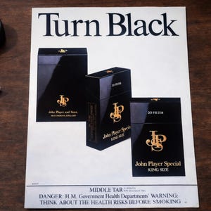 May include: An advertisement featuring three black cigarette boxes with gold lettering and logos. The text "Turn Black" is at the top. The boxes are labeled "John Player Special" and "John Player and Sons." A warning about health risks is at the bottom.