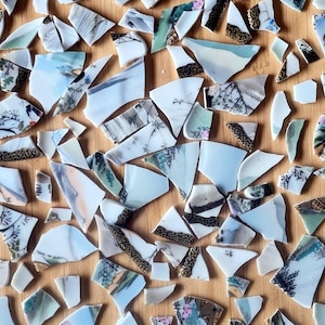 May include: An overhead view of broken porcelain shards scattered on a wooden surface. The fragments feature diverse designs, such as floral patterns, landscapes, and geometric borders. The colour palette includes shades of blue, white, brown, and gold.