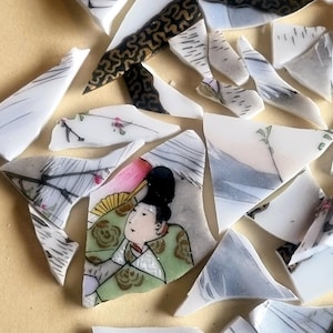 May include: A collection of broken ceramic plate fragments. The pieces feature a woman in traditional Japanese dress, floral motifs, and geometric patterns in black and gold. The fragments are scattered on a light tan surface.