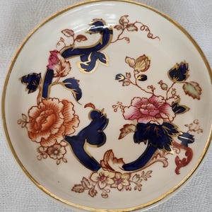 May include: A small, round, off-white dish with a gold rim. The dish is decorated with a floral pattern featuring orange, pink, and blue flowers with gold accents. The design is symmetrical and covers most of the dish's surface.