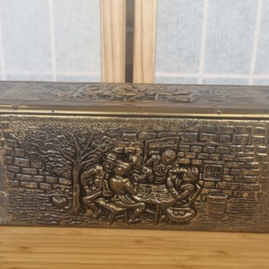 May include: An antique brass-effect box with a detailed relief scene of people around a table. The box has a textured surface and a hinged lid. The relief depicts figures, a tree, and a brick wall. The box is rectangular.
