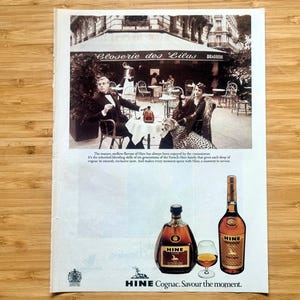 Puede incluir: Anuncio vintage de coñac HINE. La imagen presenta una escena en tonos sepia de personas en un café y dos botellas de coñac con una copa. El texto dice "HINE Cognac. Savour the moment."
