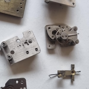 May include: A collection of disassembled watch parts, including metal plates, gears, and screws. The components are in various shapes and sizes, with a mix of silver and brass tones. A small, red jewel is also visible.