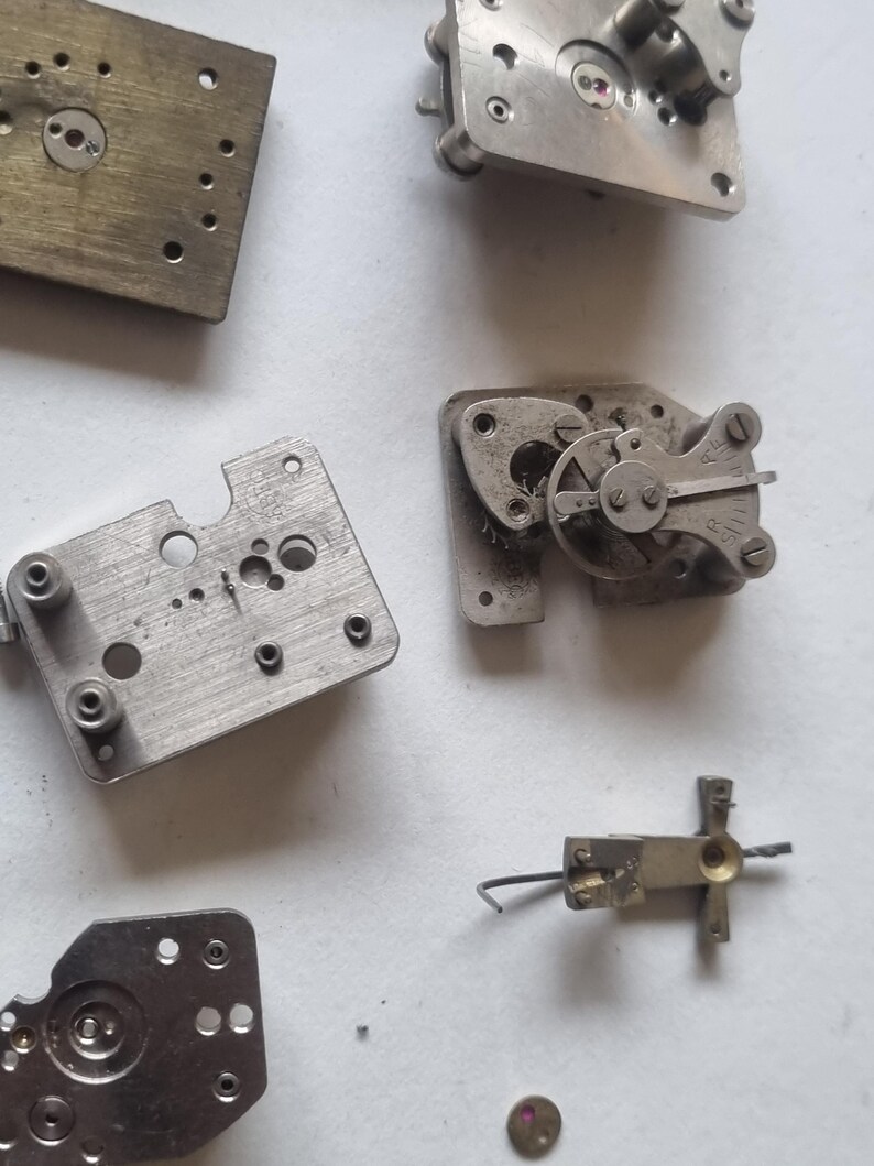 May include: A collection of disassembled watch parts, including metal plates, gears, and screws. The components are in various shapes and sizes, with a mix of silver and brass tones. A small, red jewel is also visible.