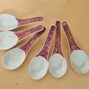 May include: Set of six white porcelain soup spoons with red and gold floral designs. The spoons have a rounded bowl and a long handle.