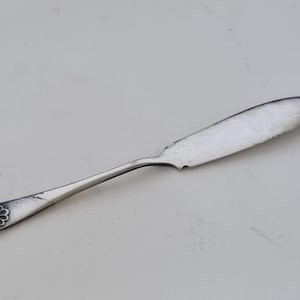 Op de afbeelding: Een zilveren botermes met een afgerond lemmet en een decoratief handvat. Het handvat heeft een schelpachtig ontwerp. Het mes is ontworpen voor het smeren van boter of andere smeersels.