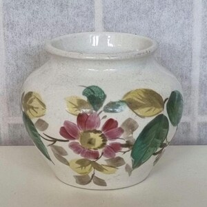 Puede incluir: Un jarrón de cerámica blanca con un diseño floral. El jarrón tiene un cuerpo redondo y una boca ancha. El diseño floral presenta flores y hojas rosas, amarillas y verdes.