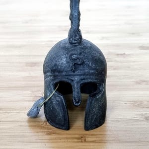 May include: A dark grey miniature Spartan helmet with a crest and face guard. The helmet has a weathered appearance and is set against a light brown wooden surface. A small, gold-coloured wire is attached to the helmet.