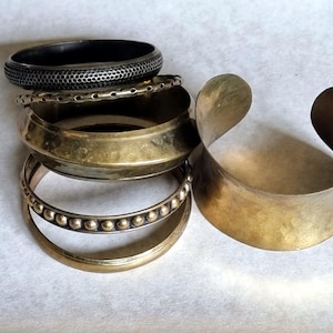 May include: A selection of brass and black metal bracelets. The bracelets include a wide cuff, a textured black band, and bands with beaded and perforated designs. The bracelets are stacked on a light-coloured surface.