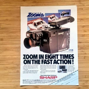 May include: An advertisement for a Sharp Super Zoom 8x camcorder. The image features the camcorder with a large lens, a racing car graphic, and the text "ZOOM IN EIGHT TIMES ON THE FAST ACTION!".