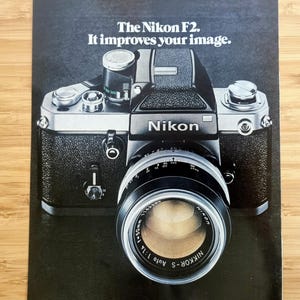 Op de afbeelding: Een vintage Nikon F2 camera met een Nikkor-S Auto 1:1.4 f=50mm lens. De camera is zwart en zilver, met het Nikon-logo prominent weergegeven. De tekst "The Nikon F2. It improves your image." staat bovenaan.