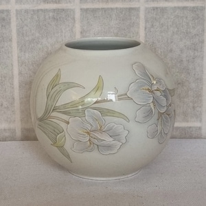 Puede incluir: Un jarrón redondo de cerámica blanquecina con un diseño floral. El jarrón presenta flores de iris azul claro y hojas verdes. El jarrón tiene un acabado liso y brillante y un interior gris.