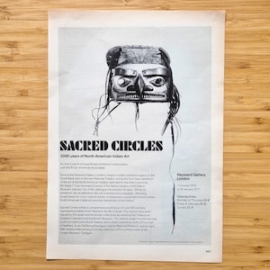 May include: A black and white poster with the text "SACRED CIRCLES" and "2000 years of North American Indian Art". The poster features a stylized illustration of a Native American mask. The poster is on a wooden surface.