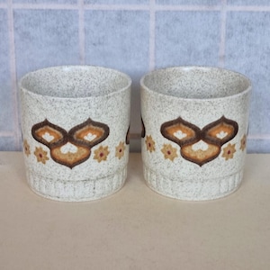 May include: Two white ceramic mugs with a brown, orange, and white geometric pattern. The mugs have a speckled texture.
