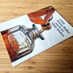 May include: Vintage advertisement for Johnnie Walker whisky. The image features a detailed illustration of a whisky bottle and a glass being filled. The text reads "a large Johnnie Walker...strictly for whisky drinkers."