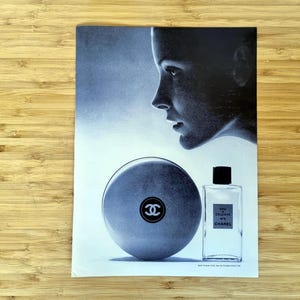 Original 1960s Chanel No. 5 magazine advertisement showing a minimalist composition with a softly lit female profile alongside the iconic round bath powder container and rectangular perfume bottle. A refined black-and-white luxury beauty image reflecting mid-century French fashion and fragrance advertising.