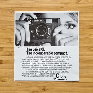 May include: Black and white advertisement for the Leica CL camera. The image shows a woman holding the camera up to her eye. The text reads "The Leica CL. The incomparable compact."