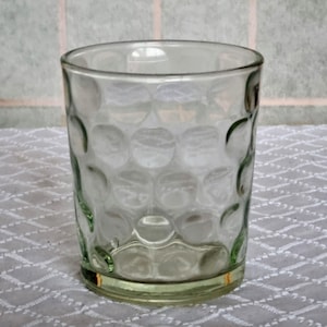 Può includere: Bicchiere trasparente con un motivo strutturato a forma di bolle. Il bicchiere ha una base e un bordo leggermente tinti di verde. È appoggiato su una superficie bianca a motivi, che suggerisce un'ambientazione informale.