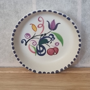 Könnte beinhalten: Cremefarbene Keramikplatte mit handgemaltem Blumendesign. Das Design zeigt lila, rosa, gelbe und grüne Elemente. Der Rand ist mit dunkelblauen Punkten verziert. Ein dekoratives Stück für die Küche oder das Esszimmer.